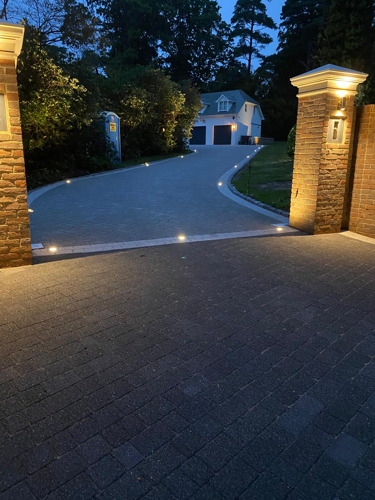 Block-paved driveway at dusk with recessed uplights and lit brick pier entrance